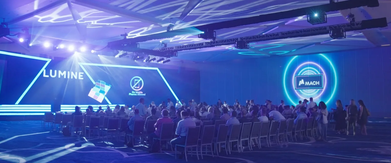 Audience seated in a ballroom facing a Lumine stage with blue LED screens and stage lights at a corporate event